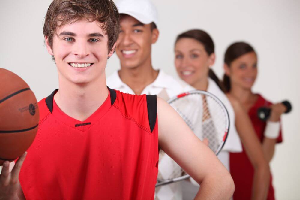 Young athletes in sports uniforms holding basketball and tennis racket, smiling at camera indoors.