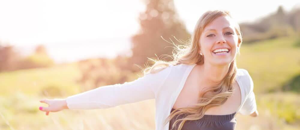 Happy woman with outstretched arms smiling outdoors in sunny field setting.