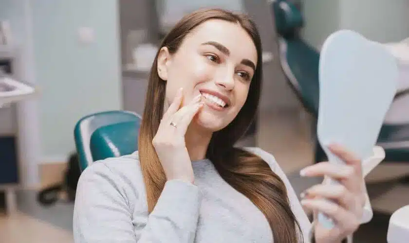 Woman admiring her smile in mirror at dental office, wearing casual clothing indoors.