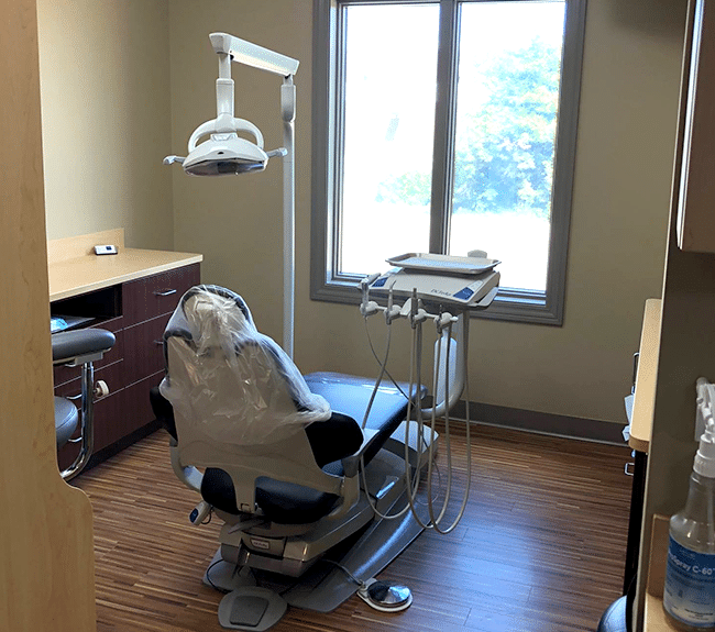 Modern dental operatory with chair, overhead light, and panoramic window overlooking nature.