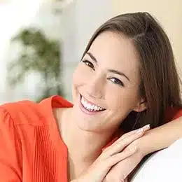 Smiling woman in orange top resting head on hand in bright, welcoming dental office setting.
