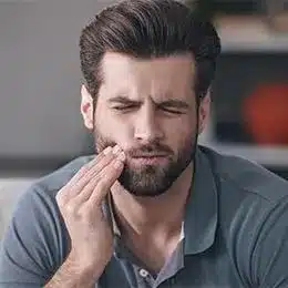 Man with beard touching cheek in pain, indoor setting, appearing to have dental discomfort