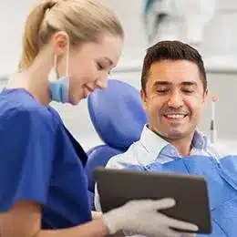 Two dental professionals in blue scrubs reviewing patient records on a tablet in a clinical setting.