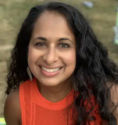 Smiling woman with long dark curly hair wearing red sleeveless top, indoor portrait setting.