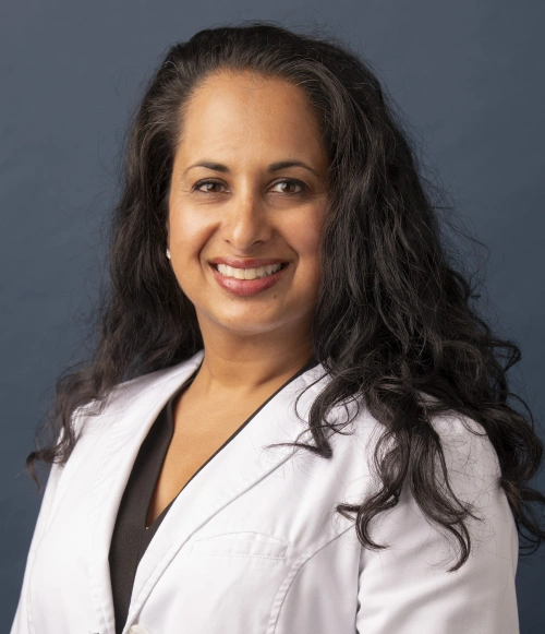 Professional headshot of woman in white dental coat against gray background, smiling at camera.